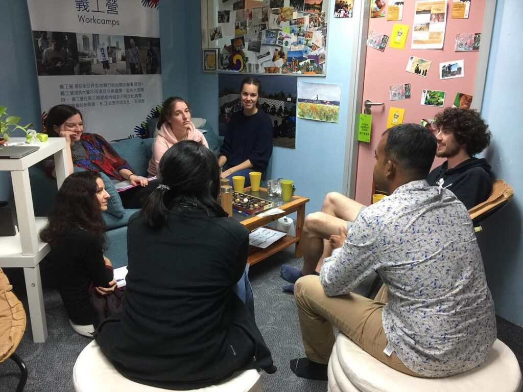 Un gruppo di persone sedute in cerchio in una stanza decorata con foto e poster. Stanno chiacchierando e giocando a un gioco da tavolo su un tavolino di legno al centro della stanza.