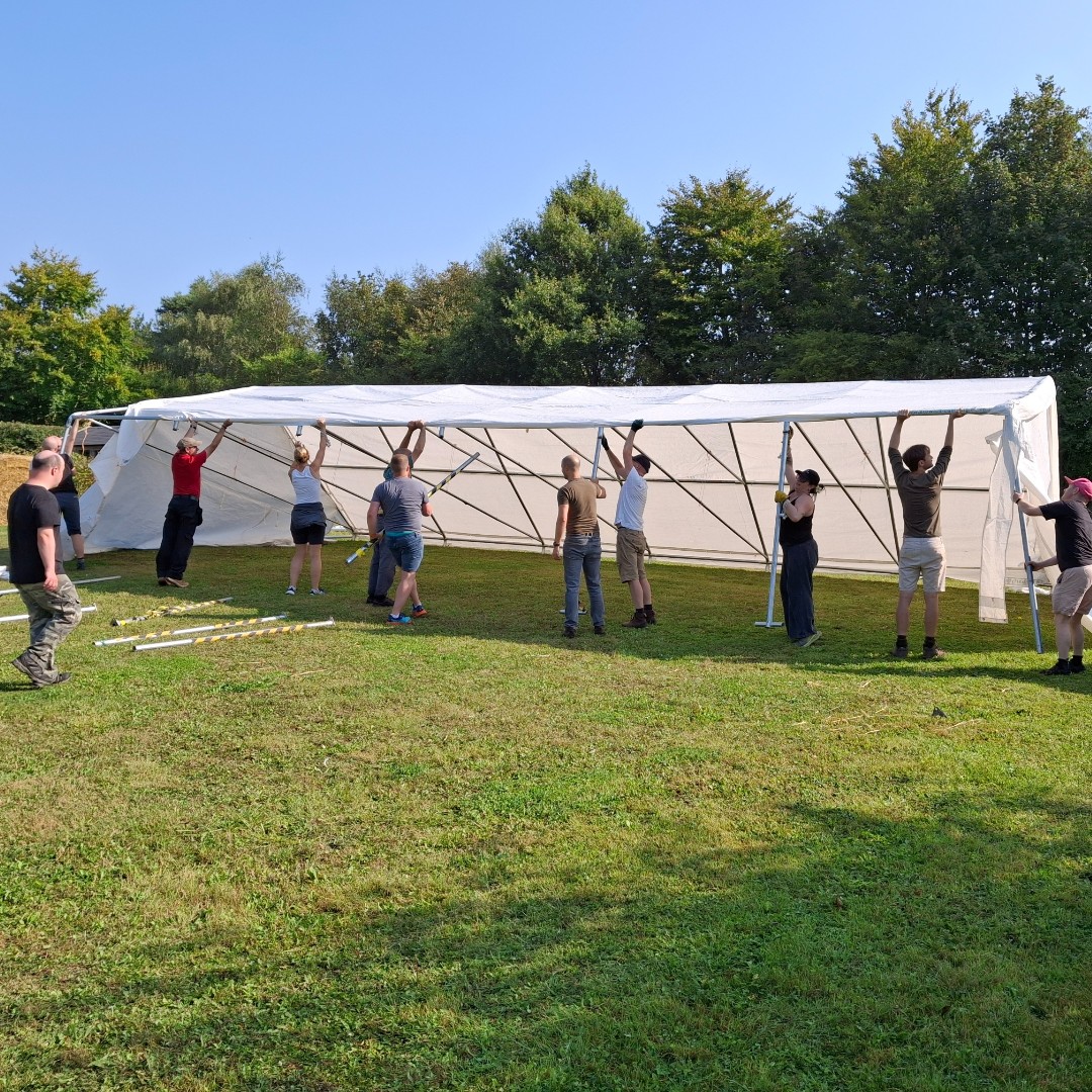 Un gruppo di persone sta montando una grande tenda bianca su un prato verde, lavorando insieme per sollevare la struttura metallica sotto il sole.